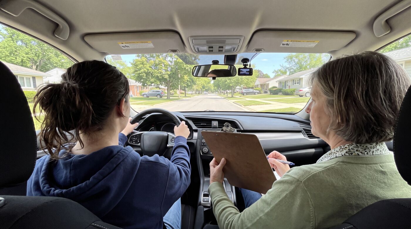 A behind-the-wheel driving lesson with a student at the wheel and an instructor beside them on a quiet suburban street